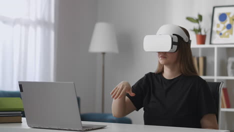 young-female-user-is-viewing-screen-of-laptop-by-modern-vr-head-mounted-display-gesticulating-by-hands-for-control-medium-female-portrait-at-home